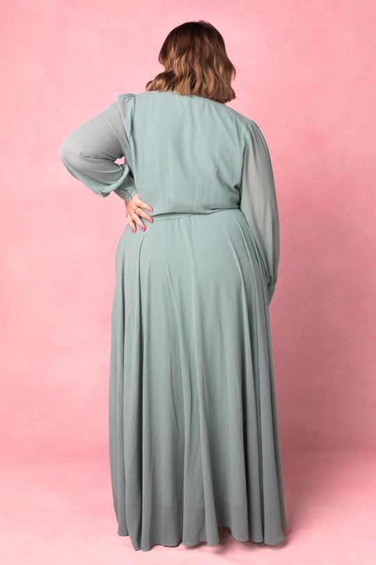 A woman stands with her back to the camera, wearing the Andie Dress in Sage by Ivy City Co—a long, flowing maxi dress showcased against a pink background. Her right hand rests on her hip, emphasizing the elegant silhouette that makes this gown an ideal choice for bridesmaids or any special occasion.