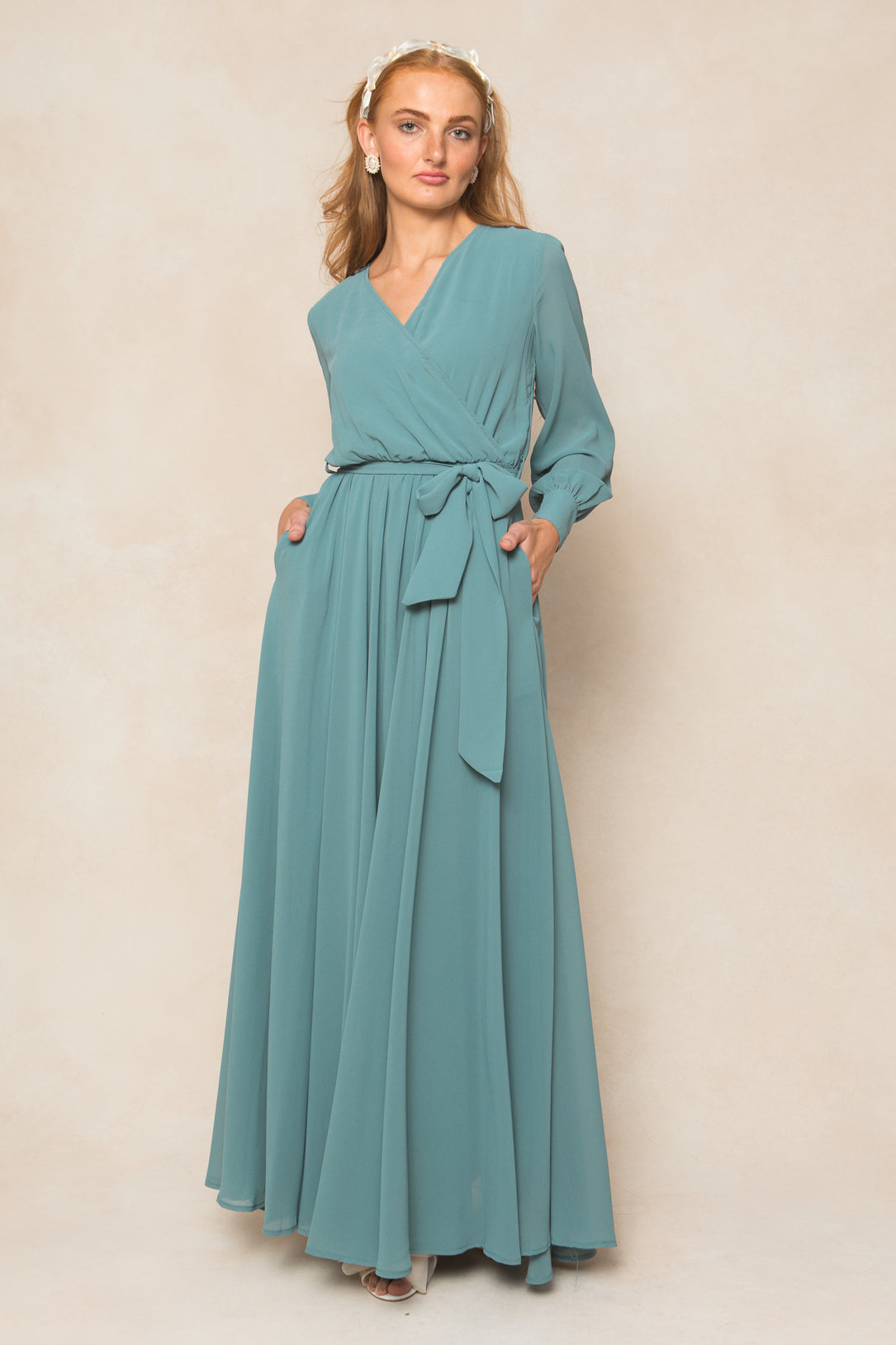 A woman in Ivy City Co’s Andie Dress in Seaglass Chiffon—a long-sleeved, waist-tied maxi—stands against a beige background, accessorized with pearl earrings and a matching headband, her hands tucked into the dress pockets.