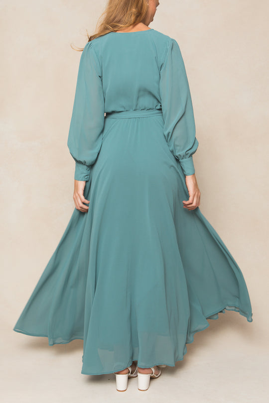 A woman with long hair stands with her back to the camera in the Ivy City Co Andie Dress in Seaglass Chiffon, featuring long sleeves and a belted waist. She pairs it with white heels against a neutral background—ideal for fall bridesmaids.