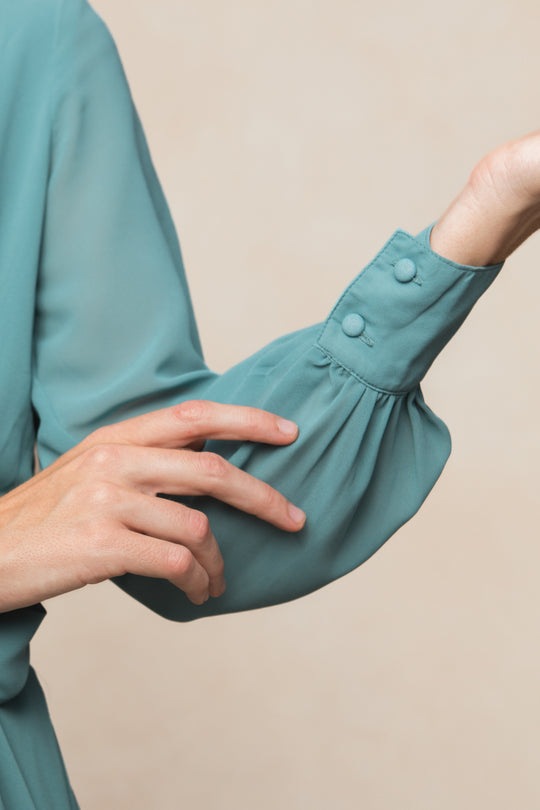 A person in the Ivy City Co Andie Dress in Seaglass Chiffon gently touches their sleeve, capturing the dress's elegance against a neutral beige background.