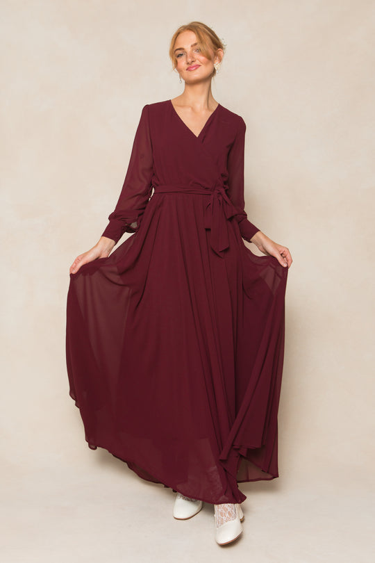 A woman wearing the Andie Dress in Wine Chiffon by Ivy City Co poses against a neutral background, showing off the long sleeves and tie waist. She styles her blond hair in a loose updo and pairs the maxi dress with white boots.