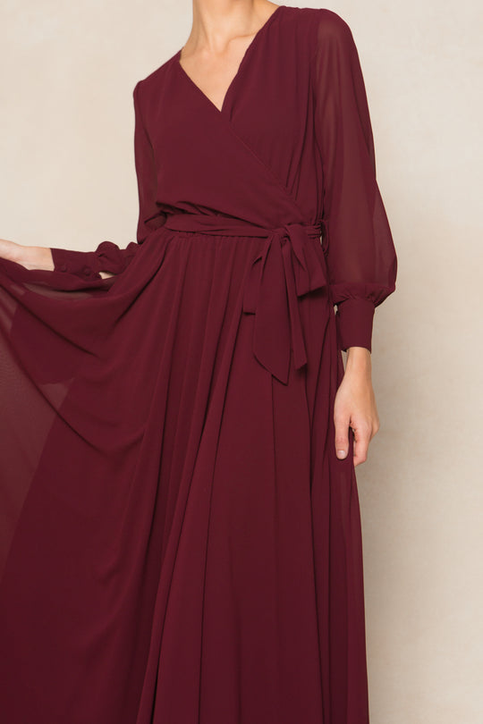 A person models the Ivy City Co Andie Dress in Wine Chiffon, featuring long sleeves and a tied waist belt, holding out one side of the flowing maxi skirt. The face is not visible.