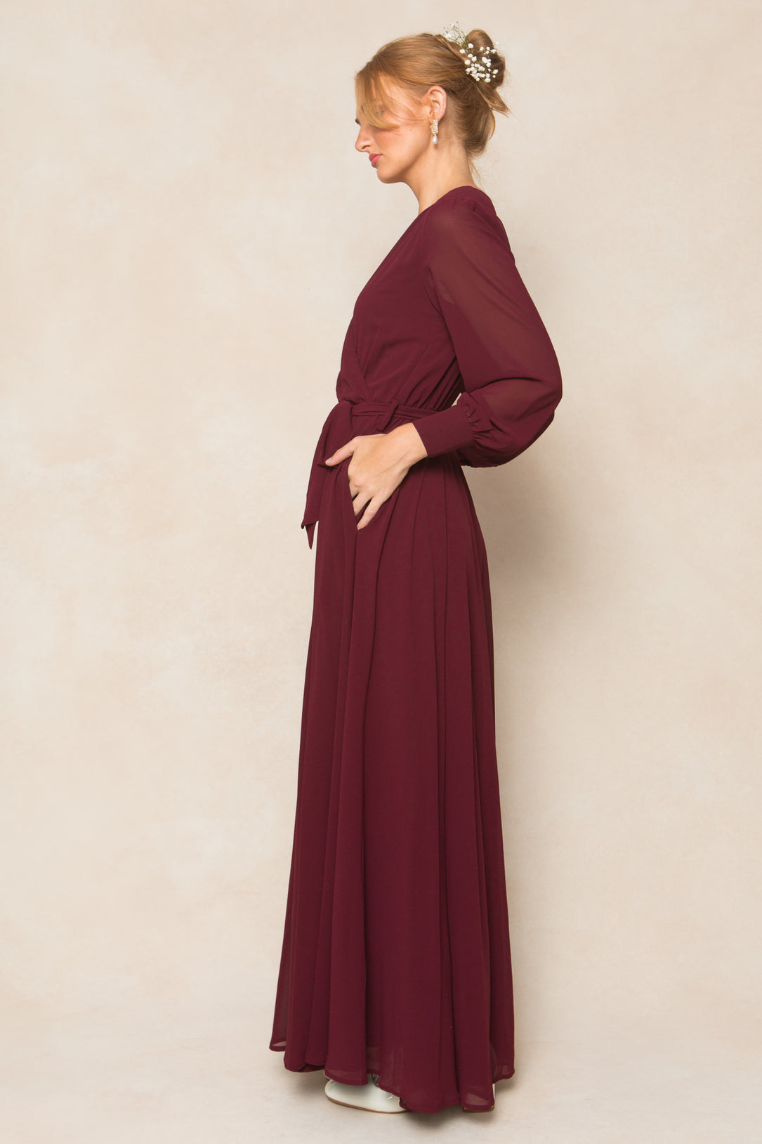 A woman in profile models the Ivy City Co Andie Dress in Wine Chiffon—a long-sleeved, floor-length gown with a tied waist—her hair styled in an updo with a decorative hairpin, set against a neutral background.