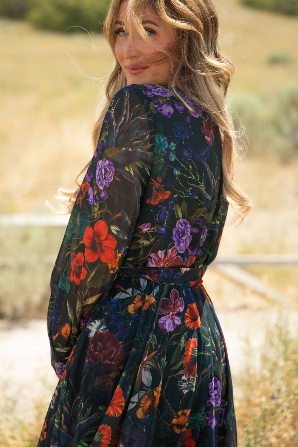 A woman with wavy blonde hair stands outdoors in a sunlit, grassy field, looking back over her shoulder and smiling. She wears the Ivy City Co Andie Dress in Evergreen Vivid Meadow—a sheer chiffon maxi dress decorated with a vibrant floral print.