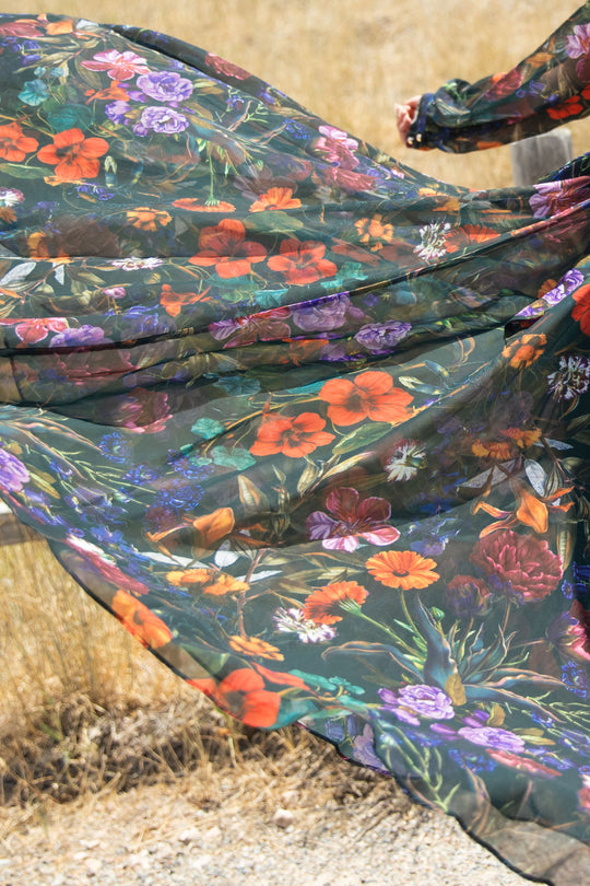 The Andie Dress in Evergreen Vivid Meadow by Ivy City Co, a flowing, semi-transparent chiffon maxi with a vivid floral pattern, is showcased outdoors against a backdrop of dry grass and a wooden fence.