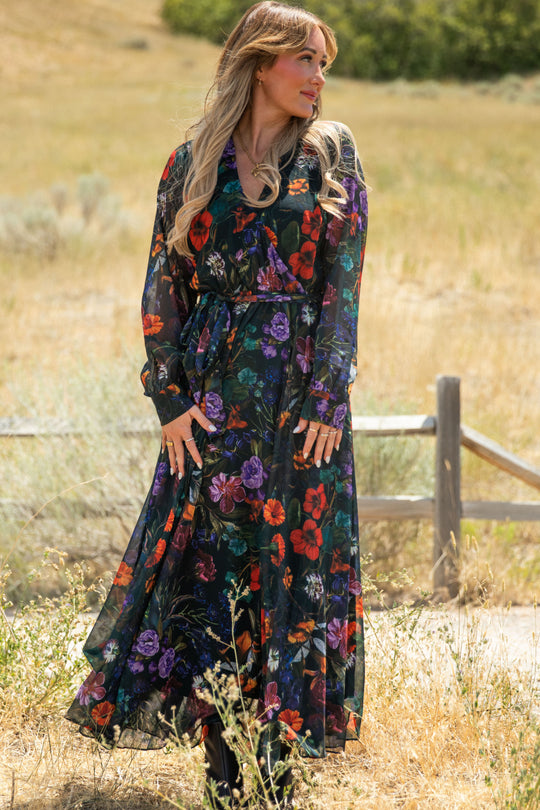 A woman stands outdoors in a field, wearing the Ivy City Co Andie Dress in Evergreen Vivid Meadow—a long, sheer floral chiffon maxi dress featuring vibrant red, purple, and green patterns. She has long, wavy blonde hair and looks to the side, with a wooden fence and dry grass visible in the background.