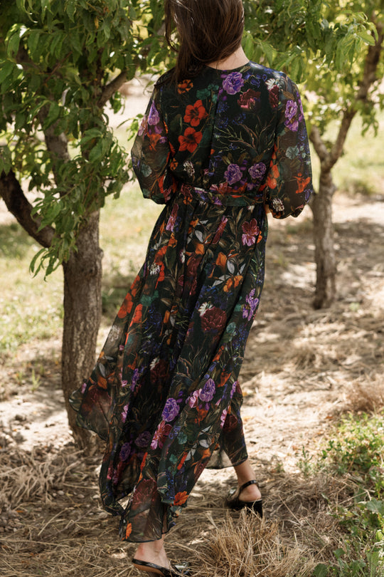 A woman with loose brown hair walks outdoors among trees on a sunny day, her back to the camera, wearing the Ivy City Co "Andie Dress in Evergreen Vivid Meadow," a long, sheer chiffon maxi dress with a vibrant floral print.