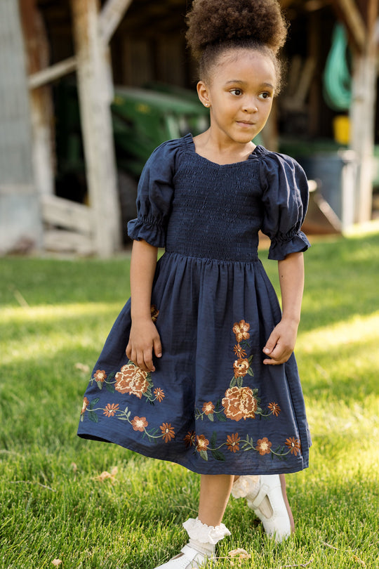 A young girl with an afro hairstyle stands on the grass, looking to the side, wearing the Ivy City Co Mini Teagan Dress in Navy Embroidered Fall Floral. The dress features balloon sleeves and delicate floral embroidery. She pairs it with white shoes and frilly socks, while a rustic wooden barn appears in the background.