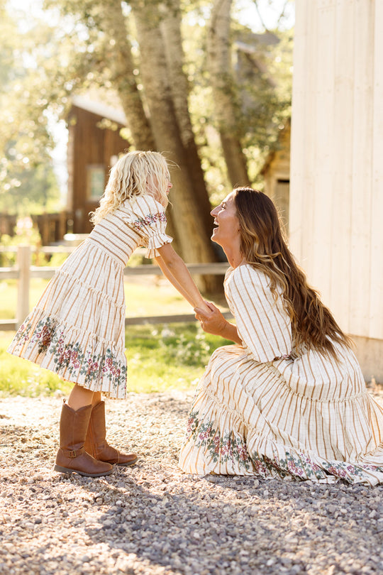 A woman kneels, smiling as she holds hands with a laughing young girl. Both wear matching Ivy City Co Mini Madeline Dresses in Embroidered Cider Grid, featuring smocked bodices and charming puff sleeves. Sunlight filters through the trees in a rustic outdoor setting.