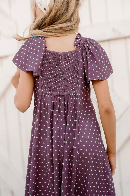 A girl with blonde hair, wearing the Ivy City Co Teen Blakely Dress in Aubergine Ditsy Dot, stands with her back to the camera in front of a white wooden fence.