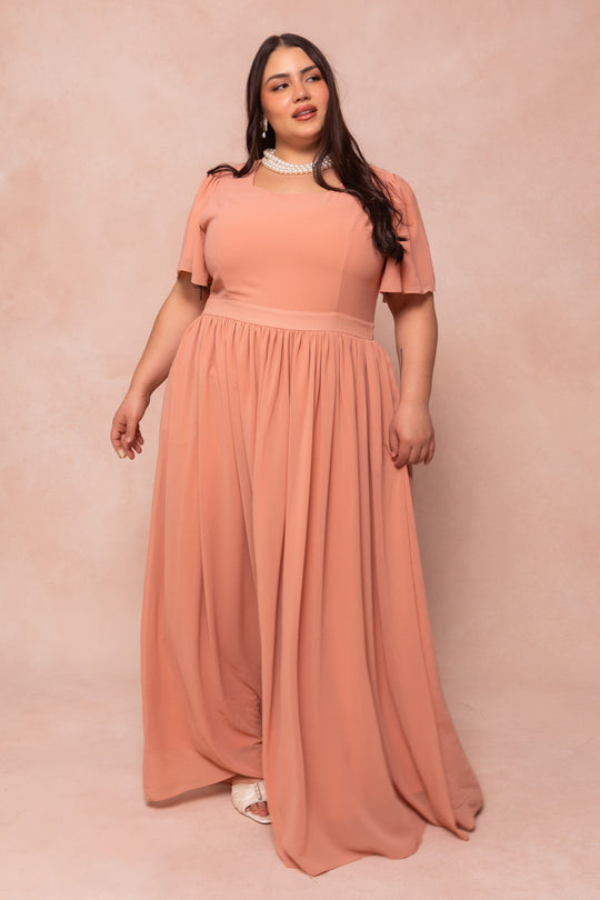 A woman in the Abbie Dress in Apricot Crush by Ivy City Co stands against a beige background, accessorized with a pearl necklace and white sandals—an elegant pick for bridesmaids or special occasions.