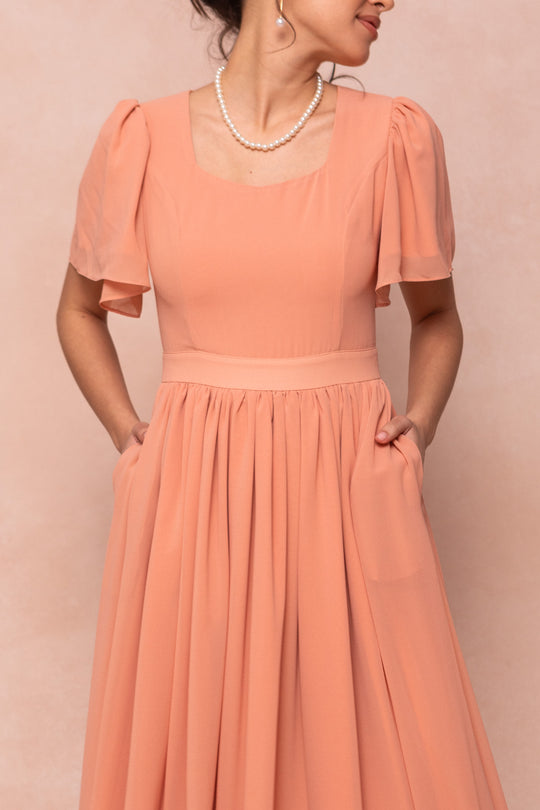 A woman wears the Abbie Dress in Apricot Crush by Ivy City Co, featuring short puffed sleeves and a square neckline, accessorized with a pearl necklace—an elegant choice for bridesmaids against a soft pink background.