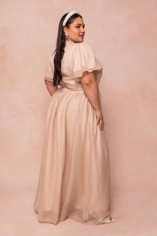A woman with long brown hair wears the Ivy City Co Abbie Dress in Champagne, styled with a pearl headband and earrings. She poses against a beige background, looking over her shoulder with a slight smile.