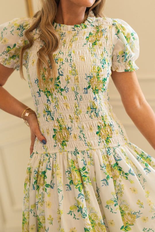 A woman wearing the Ivy City Co Jennifer Dress in Garden Tea Party Floral—a short-sleeved, smocked dress featuring yellow and green floral prints on lightweight fabric—poses indoors with one hand on her hip, showing only her torso and part of her face.