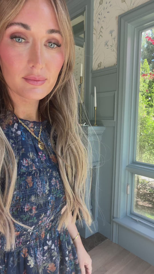 A woman with long, wavy blonde hair stands indoors by a window, framed by greenery and illuminated by soft natural light. She is wearing the Ivy City Co Leonora Dress in Midnight Autumn Bouquet—a blue floral dress featuring puff sleeves and a maternity-friendly silhouette that adds effortless charm to her look. Decorative candles are visible in the background.
