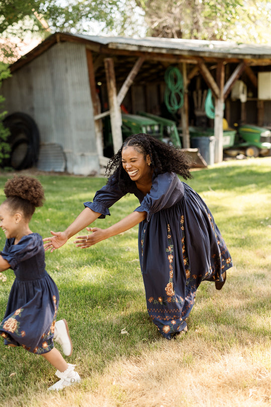A woman and a young girl, both wearing the Ivy City Co Mini Teagan Dress in Navy Embroidered Fall Floral—with balloon sleeves and smocked bodices—play and run on the grass near a rustic shed surrounded by farm equipment. The woman smiles and reaches out as the girl runs ahead of her.