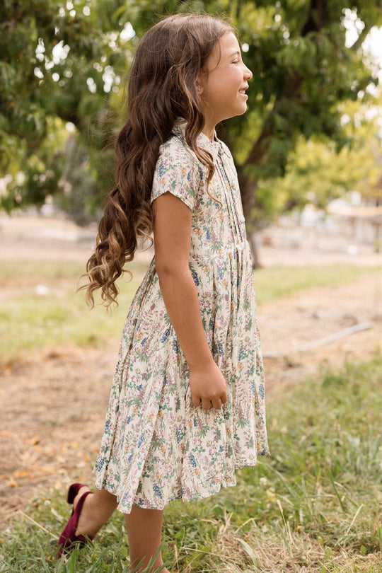 A young girl with long wavy hair stands sideways outdoors in a grassy area with trees in the background, smiling while wearing the Ivy City Co Mini Amber Dress in Autumn Bouquet.
