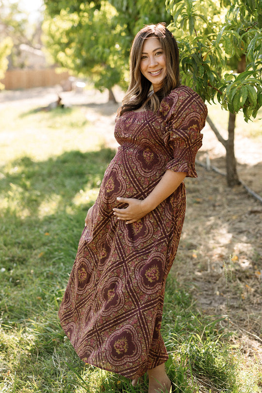 Wearing the Ivy City Co Josephine Dress in Nocturne Diamond Rose—a patterned cotton dress with a smocked bodice—a pregnant woman smiles and stands outdoors, holding her belly with one hand. The maternity-friendly dress flows comfortably as she stands near a green tree on a sunny day.