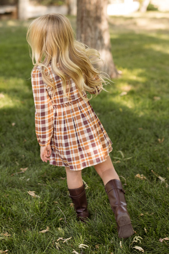 A young girl with long blonde hair, wearing the Ivy City Co Mini Bowie Long Sleeve Dress in Spiced Plaid and brown boots, walks on green grass near a tree with her back to the camera.