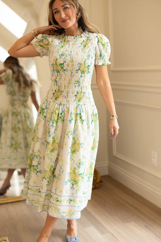 A woman smiles as she stands on a wooden floor in a well-lit room, wearing the Ivy City Co Jennifer Dress in Garden Tea Party Floral. This white floral print dress features short puff sleeves, is crafted from lightweight fabric, and is designed to be maternity friendly. The mirror behind her reveals both the front and back views of the dress.
