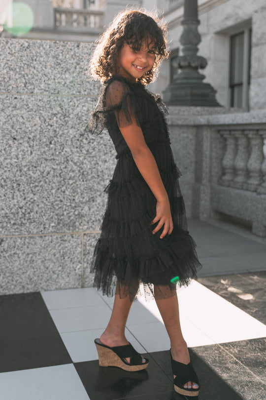A young girl wearing the Ivy City Co Mini Whimsical Dress in Black, featuring a layered tulle skirt, poses and smiles outdoors on a checkered floor with sunlight shining on her, styled with wedge sandals.
