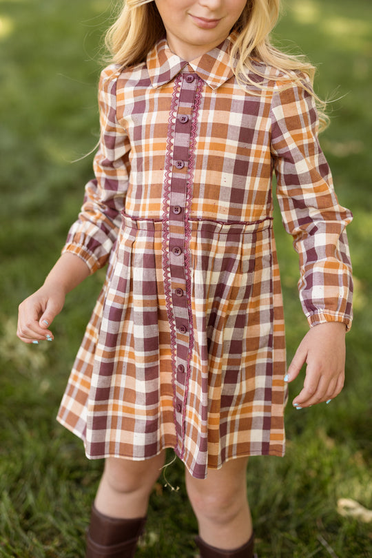 A young girl stands on grass wearing the Ivy City Co Mini Bowie Long Sleeve Dress in Spiced Plaid, a cotton dress in shades of brown, orange, and cream with charming balloon sleeves, front buttons, and decorative trim. Only her torso and legs are visible.