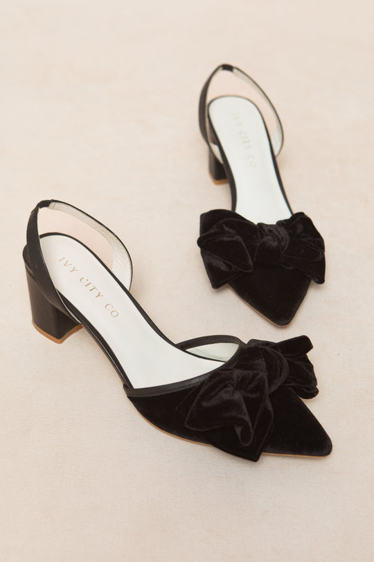 The Eloise Bow Heel in Black by Ivy City Co features black velvet slingbacks with pointed toes, low block heels, large velvet bows on the toes, and a white branded insole, displayed on a light beige surface.