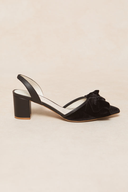 The Eloise Bow Heel in Black by Ivy City Co is a black velvet slingback with a closed pointed toe, large front bow detail, and mid-height block heel, shown against a light beige background.