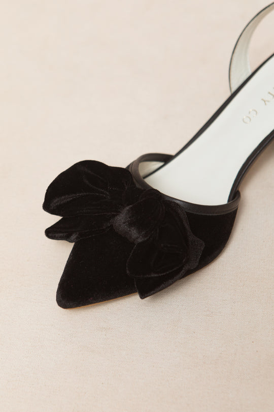 A close-up of the Ivy City Co Eloise Bow Heel in Black, a black velvet pointed-toe shoe with a large matching bow on the front, perfect for lovers of elegant black heels, displayed on a light beige surface.