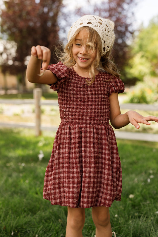 A young girl stands on the grass outdoors with trees in the background, smiling and holding something small in her hand. She is wearing the Ivy City Co Mini Tori Dress in Cranberry Popcorn Plaid with puff sleeves, paired with a white crocheted hat.