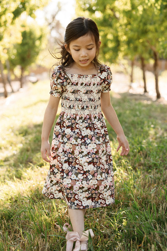 A young girl in the Ivy City Co Mini Sunday Best Dress in Midnight Garden, featuring a smocked bodice and puff sleeves, walks outdoors along a grassy path surrounded by green trees as sunlight filters through the leaves.