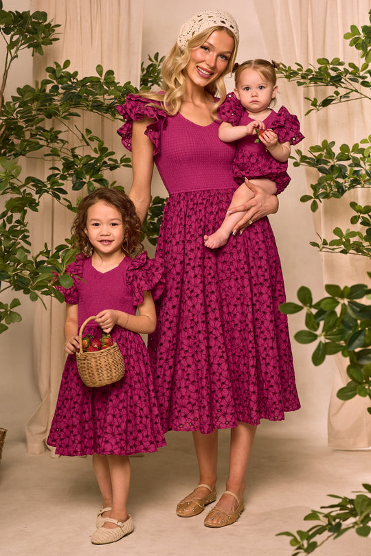 A woman and two girls wear matching Ivy City Co Hattie Dresses in Magenta with ruffled sleeves and lace details. The older girl holds a small basket, set against green plants and beige curtains for a charming look.