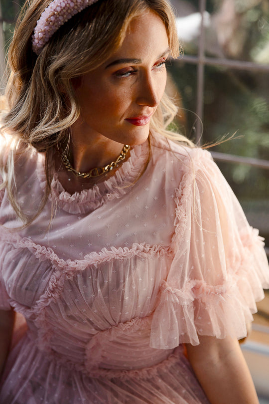 A woman in Ivy City Co’s Whimsical Dress in Blush, featuring sheer ruffles and puffed sleeves, pairs it with a textured headband and gold necklace. Sunlight highlights her wavy blonde hair as she gazes to the side.