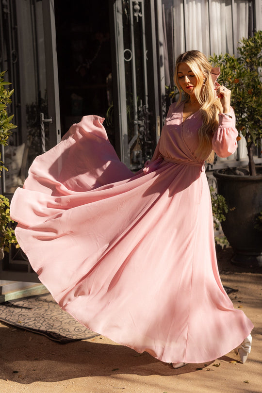 A woman twirls outdoors in the sunlight, the skirt of her Ivy City Co Andie Dress in Blush flowing gracefully. She stands by an open door framed by plants, her long blonde hair cascading loosely over her shoulder.