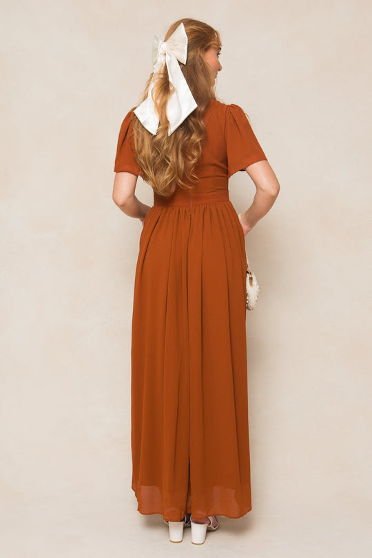 A woman stands with her back to the camera, wearing the Ivy City Co Abbie Dress in Amber Chiffon—a flowing maxi-length gown elegantly styled with white platform heels and a large white bow adorning her long wavy hair. In her left hand, she carries a small white bag.