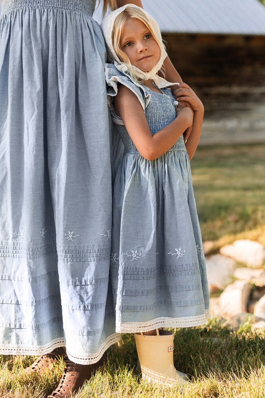A young blonde girl in the Ivy City Co Hattie Ankle Length Dress in Embroidered Chambray, featuring a smocked bodice, hugs an adult wearing a matching dress. They stand on grass near a rustic building. The girl, looking thoughtful and wearing beige rain boots, embodies the charm of this maternity-friendly style—perfect for creating memories together.