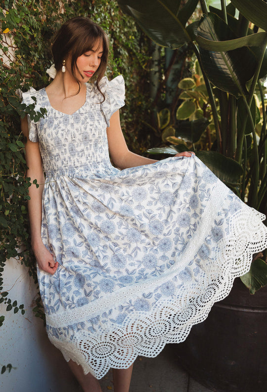 A woman in the Ivy City Co Hattie Dress in Blue Floral stands by lush greenery, gently holding out the skirt and looking down with a soft expression.