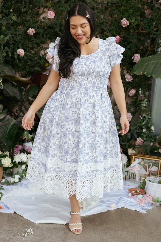 A woman with long dark hair smiles outdoors on a picnic blanket, wearing the Ivy City Co Hattie Dress in Blue Floral, surrounded by flowers and greenery.