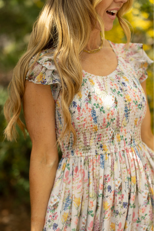 A woman with long, wavy blond hair smiles outdoors in soft sunlight while wearing the Ivy City Co Hattie Dress in Cordelia Floral, a maternity-friendly A-line midi. Lush green and yellow foliage appear blurred in the background.