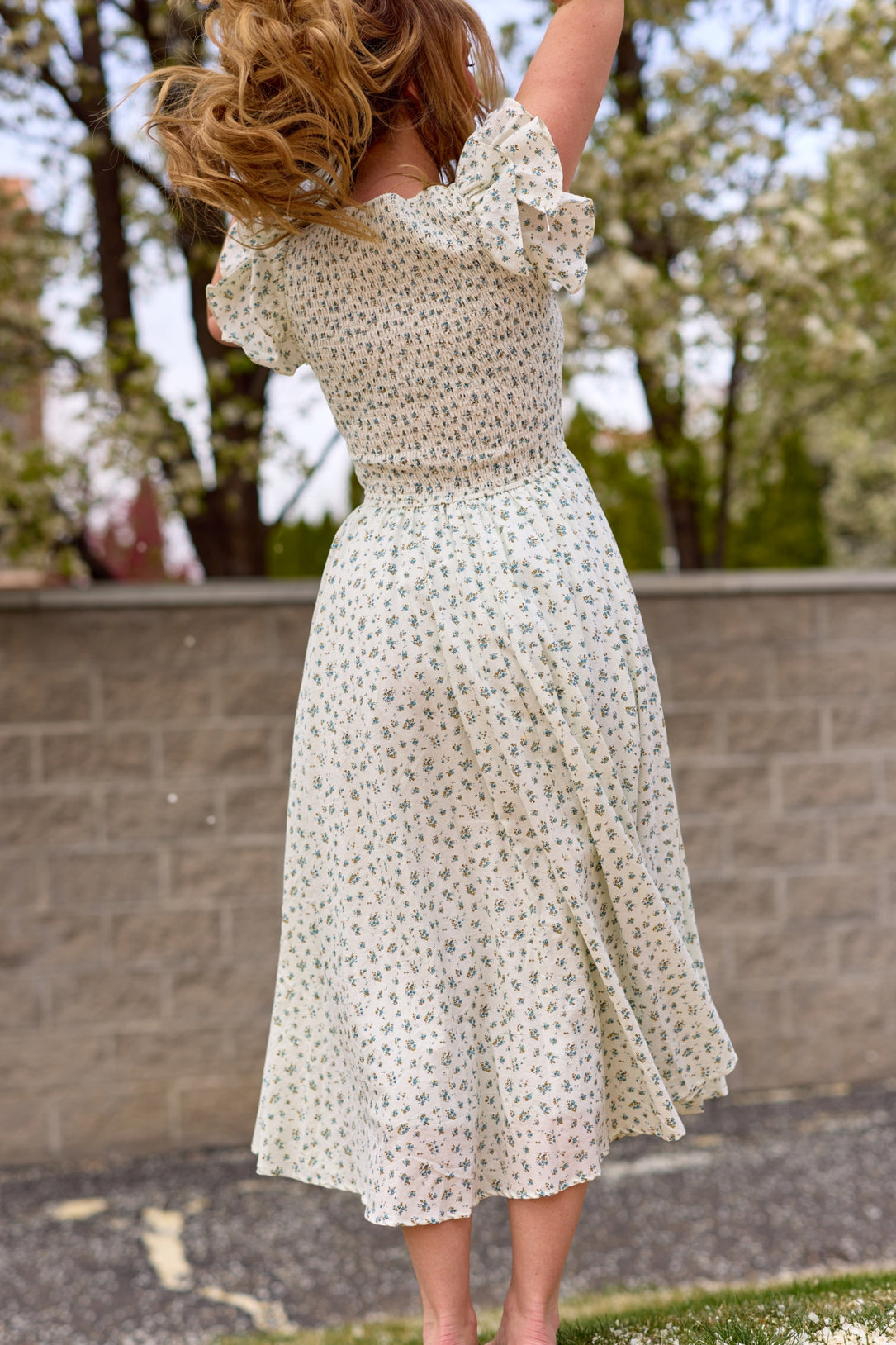 A woman jumps with her back to the camera, hair airborne, wearing the Ivy City Co Hattie Dress in Madeline Blue. She stands outdoors before a stone wall, with blooming trees in the background.