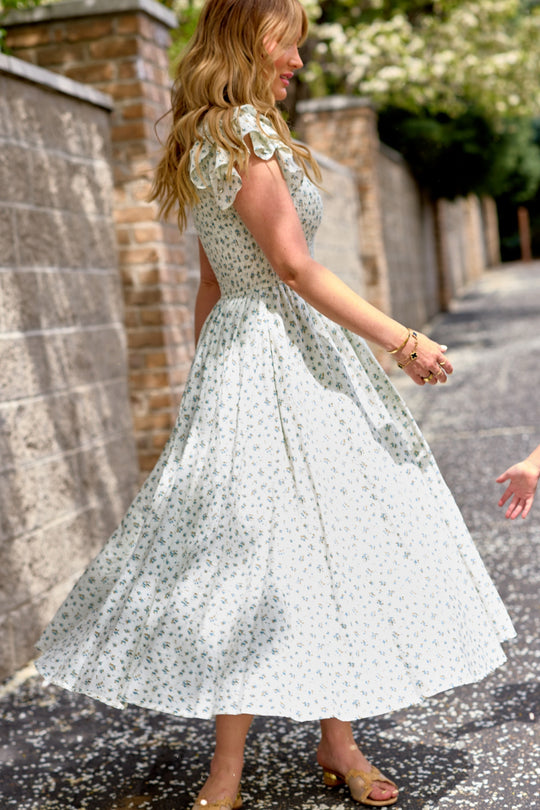 A woman twirls outdoors on a sunny day wearing the Ivy City Co Hattie Dress in Madeline Blue, her wavy blonde hair flowing and gold sandals gleaming, while a child’s hand reaches in from the right edge of the image.