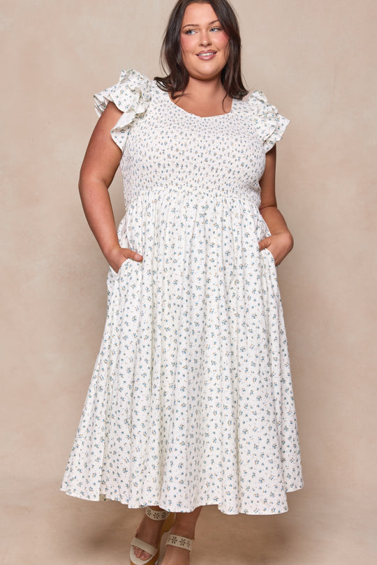A woman stands smiling with her hands in her pockets, wearing the Ivy City Co Hattie Dress in Madeline Blue—a sleeveless cotton midi dress with ruffled shoulders—paired with light-colored sandals against a beige background.