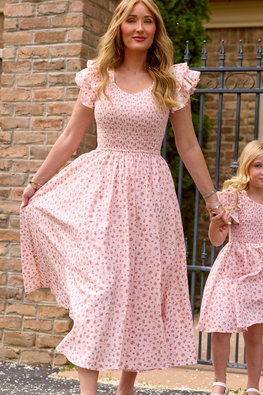 A woman in the Ivy City Co Hattie Dress in Madeline Pink holds hands with a young girl in a matching birthday dress. Both wear charming pink floral cotton midi dresses as they stand by a brick wall and iron gate.