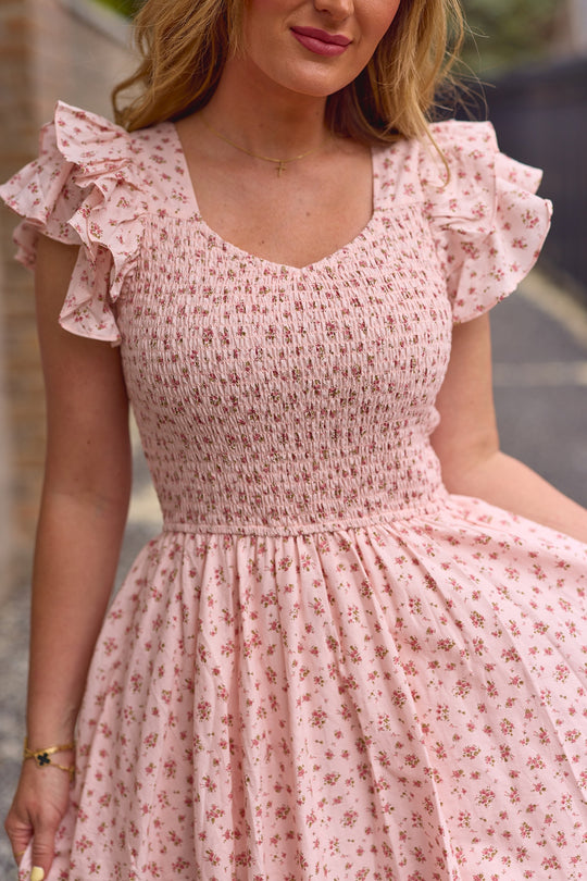 A woman stands outdoors, lightly holding the skirt of the Hattie Dress in Madeline Pink by Ivy City Co—a pink cotton midi dress with small floral prints, ruffled cap sleeves, and a smocked bodice. Her face is partially visible.