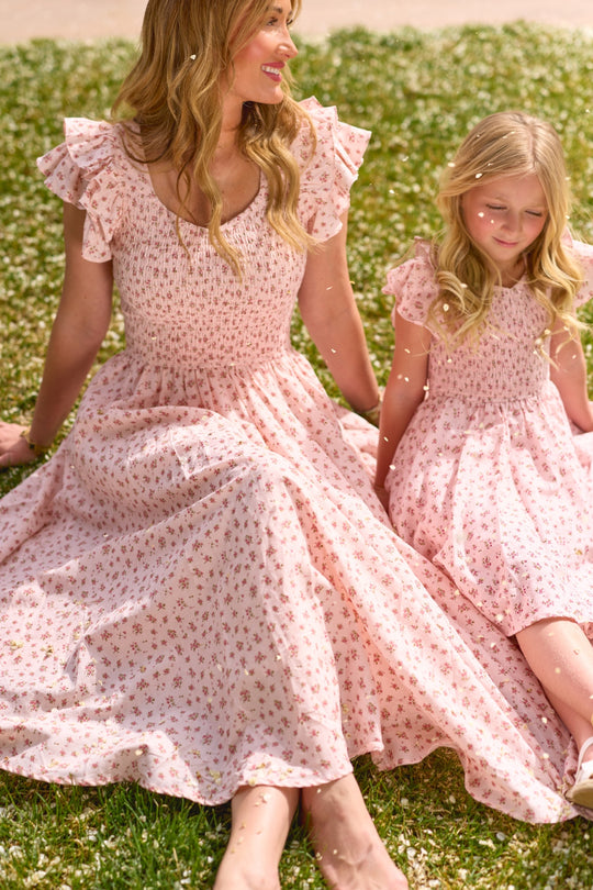 A woman and a young girl, both in Ivy City Co’s Hattie Dress in Madeline Pink, sit on grass with white flowers, smiling and enjoying a sunny day together.