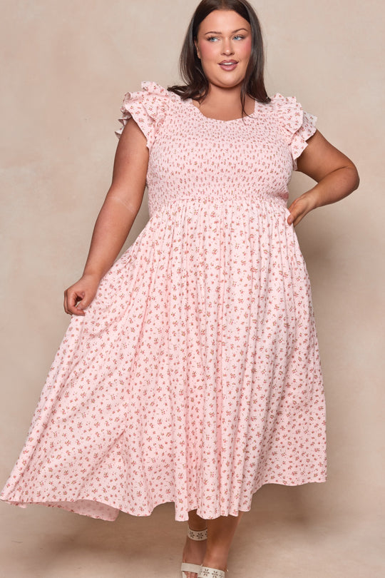 A woman in the Hattie Dress in Madeline Pink by Ivy City Co—a light pink, short-sleeved cotton midi dress with a small floral print—stands against a neutral backdrop, smiling and holding out her dress. A chic choice for a birthday girl.