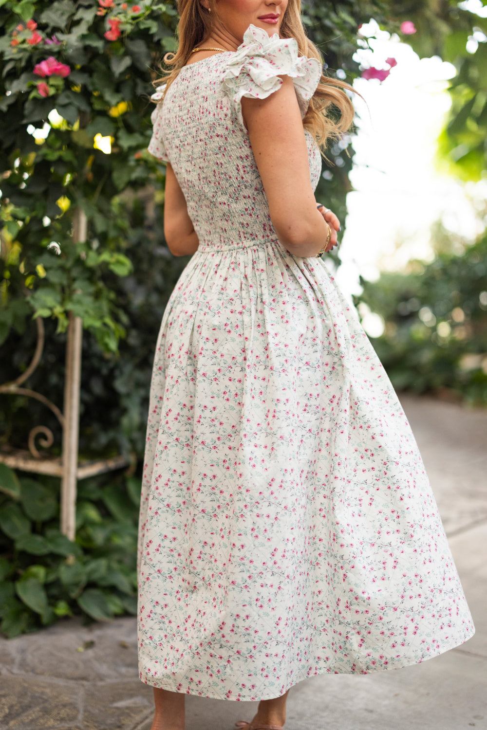 A woman stands outdoors in the Ivy City Co Hattie Dress in Pansy Trellis, a light floral cotton dress with short ruffled sleeves. She’s turned slightly to the side among greenery and flowers. This style is nursing and maternity friendly.
