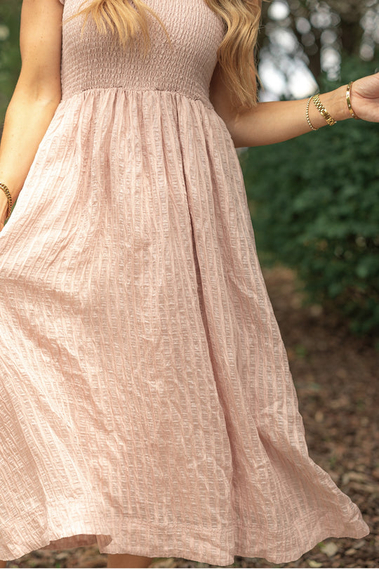 A person wears the Ivy City Co Hattie Dress in Pink Stripes outdoors, holding the skirt. Their long hair is down, gold bracelets shine on their wrist, and green foliage and trees blur softly in the background.
