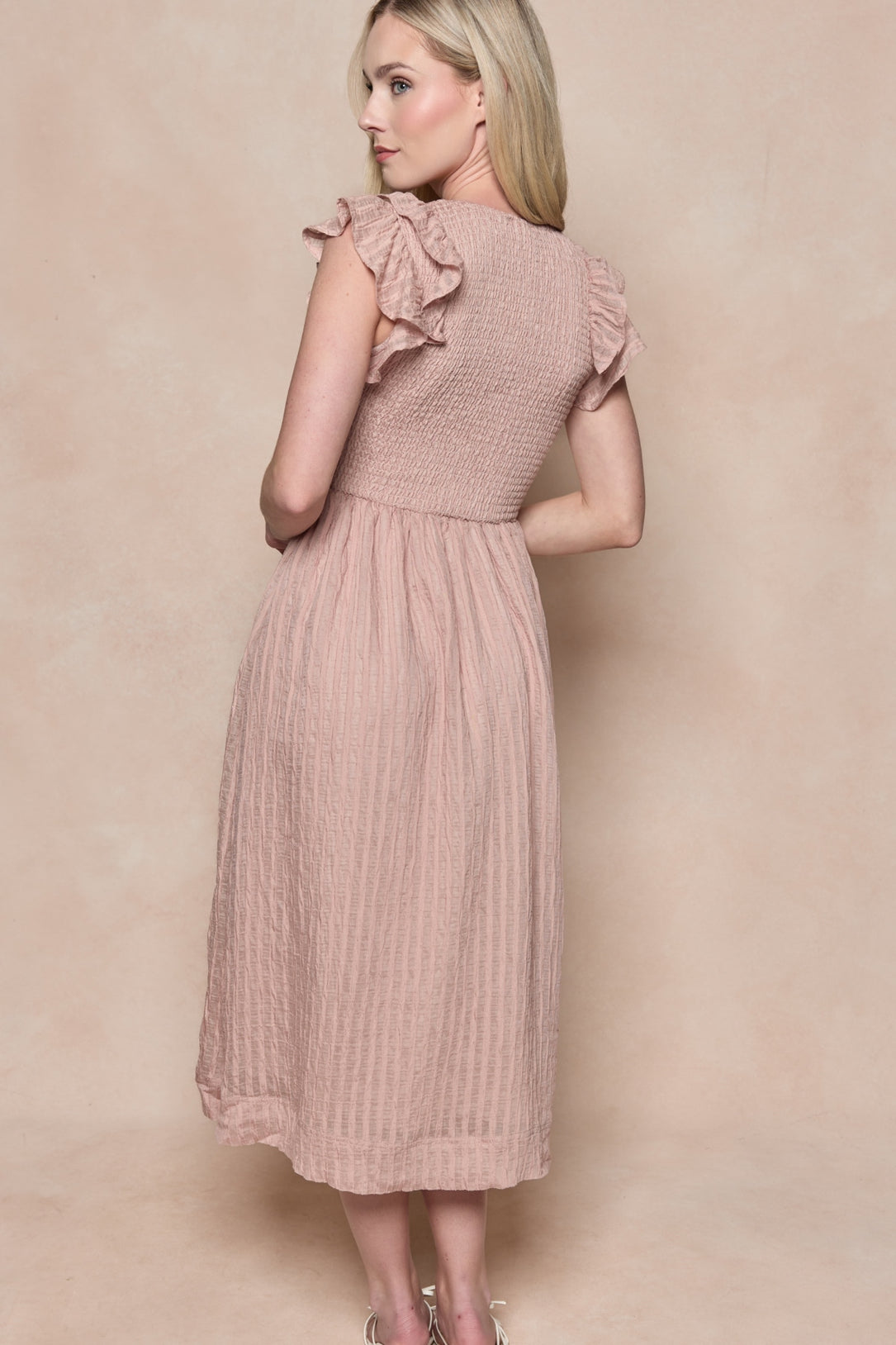 A woman with long blonde hair models the Ivy City Co Hattie Dress in Pink Stripes, featuring a smocked bodice and ruffled sleeves. The maternity-friendly midi is shown from behind as she looks over her shoulder against a beige background.