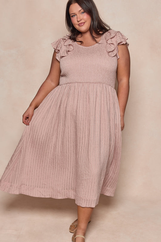 A woman smiles as she stands against a neutral background, holding one side of her Ivy City Co Hattie Dress in Pink Stripes—a sleeveless midi with smocked bodice and ruffled shoulders—paired with strappy sandals.